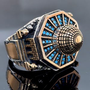 Könnte beinhalten: Ein silberner und goldener Ring mit einem detaillierten Design eines kuppelförmigen Gebäudes. Der Ring ist mit türkisfarbenen Steinen besetzt.