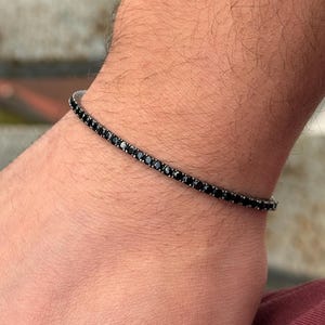 May include: A black tennis bracelet with small, round, black gemstones. The bracelet is worn on a wrist, showcasing the jewelry's design and sparkle. The bracelet is a classic piece of jewelry.