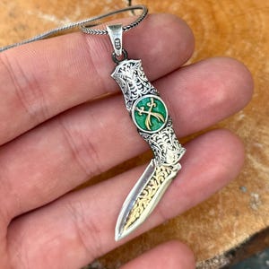 Pode incluir: Um colar com pingente de prata com um design detalhado de faca. A faca tem uma lâmina dourada com padrões ornamentados e um emblema verde e dourado. O pingente é suspenso por uma corrente de prata, mostrando intrincados trabalhos em metal.