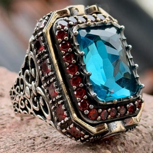 Może przedstawiać: Srebrny pierścionek z dużym niebieskim kamieniem szlachetnym otoczonym mniejszymi czerwonymi kamieniami szlachetnymi. Pierścionek ma skomplikowany wzór z złotym obrączką.