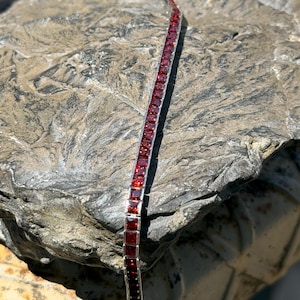 Puede incluir: Una pulsera de tenis que presenta una fila continua de piedras preciosas de granate rojo oscuro de corte cuadrado, engastadas en un canal de metal plateado pulido. La pulsera muestra un diseño uniforme con cada piedra sujeta en un marco lineal.
