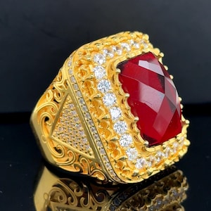 May include: A gold-colored ring with a large, faceted, red gemstone at the center. The ring is square-shaped and surrounded by smaller, clear gemstones. Intricate filigree and additional small gemstones adorn the ring's band.