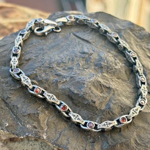 Peut inclure: Bracelet en argent avec des maillons ornés et de petites pierres précieuses rouges rondes. Le bracelet est présenté sur une surface de pierre grise texturée. Le fermoir est visible, et le design a une esthétique vintage.
