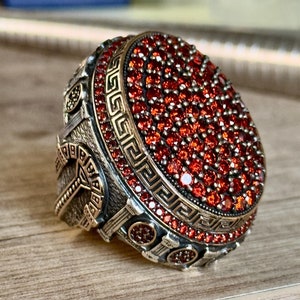 Könnte beinhalten: Ein großer, verzierter Ring mit einem silber- und goldfarbenen Metalldesign. Der Ring hat ein zentrales Oval aus zahlreichen kleinen, roten Edelsteinen. Das Band hat detaillierte Muster und einen griechischen Schlüsselrand.
