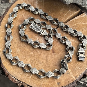 Könnte beinhalten: Eine silberne Kettenkette mit einem detaillierten, komplizierten Design. Die Kette besteht aus miteinander verbundenen, rautenförmigen Gliedern mit einem dekorativen Muster. Die Kette hat einen Verschluss.