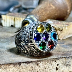 Op de afbeelding: Een zilveren ring met een ingewikkeld ontwerp en zeven kleurrijke edelstenen die in een cirkelvormige patroon zijn geplaatst. De edelstenen zijn rood, oranje, geel, groen, blauw, indigo en violet.
