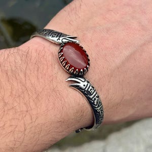 May include: A silver cuff bracelet with an oval, reddish-brown gemstone. The bracelet has an engraved design and an open, adjustable design. The gemstone is surrounded by a decorative silver setting.