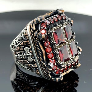 May include: A silver ring with a large, square, red gemstone surrounded by smaller red gemstones. The ring has intricate details and a textured band.