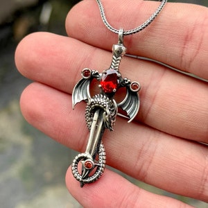 Könnte beinhalten: Silberner Anhänger mit einer detaillierten Drachenfigur, die um ein Schwert mit roten Edelstein-Akzenten gewickelt ist.