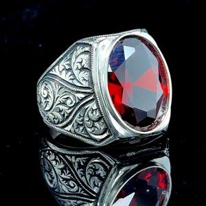 以下が含まれることがあります： 装飾的なデザインと大きな楕円形の赤い宝石が付いた銀の指輪