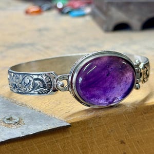 May include: A silver bracelet featuring an intricate floral design and a large, oval amethyst gemstone.