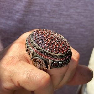 May include: A silver ring with a large, oval-shaped stone set in a decorative, ornate design. The stone is a deep red color and is surrounded by a border of smaller stones. The ring is detailed with a Greek key pattern.