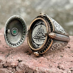 Könnte beinhalten: Ein silberner Ring mit einem Klappdeckel, der eine Inschrift in arabischer Schrift enthüllt. Der Ring ist mit komplizierten floralen Mustern und grünen Edelsteinen verziert.