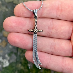 May include: A silver and gold sword pendant necklace. The sword has a curved blade and intricate details. The pendant is hanging from a silver chain.