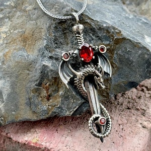 Puede incluir: Un collar con un colgante de plata que representa una espada con un dragón envuelto alrededor. La espada tiene una piedra preciosa roja en la parte superior y piedras preciosas rojas más pequeñas en los lados. El colgante está en una cadena de plata.