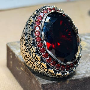 Może przedstawiać: Ozdobny pierścionek z dużym, fasetowanym, ciemnoczerwonym kamieniem. Pierścionek ma srebrną obręcz z misternymi zdobieniami filigranowymi, a centralny kamień otoczony jest mniejszymi czerwonymi kamieniami. Oprawa to połączenie srebra i złota.