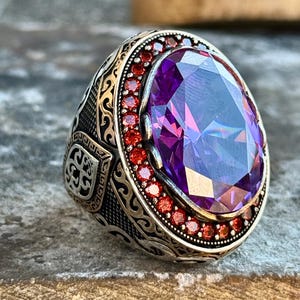 May include: A silver ring with an oval, faceted purple gemstone as the centerpiece. The ring is surrounded by small red gemstones and ornate detailing on the band. The ring is set on a textured surface.