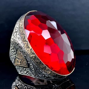 Może przedstawiać: Pierścionek z dużym, fasetowanym, czerwonym kamieniem osadzonym w ozdobnej srebrnej obrączce. Obrączka ma szczegółowe wzory kwiatowe i geometryczne ze złotymi akcentami. Pierścionek jest umieszczony na ciemnym tle, podkreślając blask kamienia.