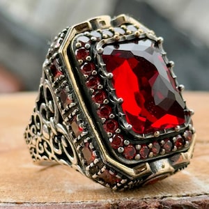 Könnte beinhalten: Ein silberner Ring mit einem großen, roten, facettierten Edelstein, der von kleineren roten Edelsteinen umgeben ist. Der Ring hat ein kompliziertes, verziertes Design.