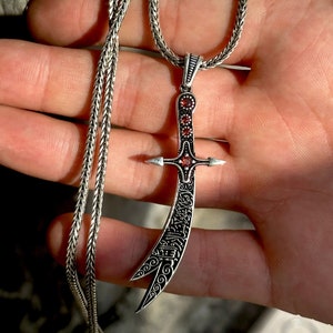 以下が含まれることがあります： 銀色のチェーンネックレスに、銀製の三日月刀の形をしたペンダントがついたもの。刀は複雑な模様と赤い宝石で飾られている。