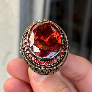 Peut inclure: Une bague ornée d'une grosse pierre rouge ovale et facettée. Le sertissage est de style ancien, avec de petites pierres rouges et des détails en métal doré et argenté. Un bijou de caractère, avec un design élaboré.