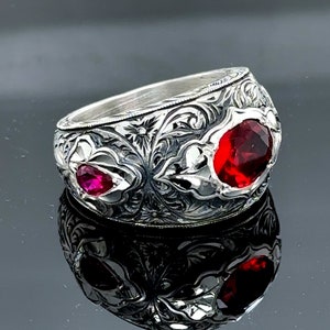 Peut inclure: Une bague en argent avec des motifs floraux complexes et deux pierres pr&eacute;cieuses rouges. La bague est large et a un profil haut.