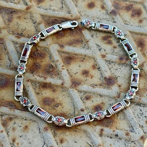 Peut inclure: Bracelet en argent orné de pierres précieuses rouges rectangulaires et de petites pierres rouges. Le bracelet présente un design décoratif et une fermeture à fermoir. Le bijou est présenté sur une surface métallique texturée.