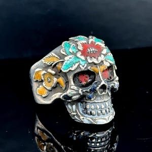 May include: A silver skull ring with colorful floral accents. The skull features red eyes and is adorned with a red, turquoise, and yellow flower design on the forehead. The ring has a detailed, gothic aesthetic.