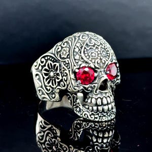 Pode incluir: Um anel de caveira prateado com intrincados detalhes florais e de rolagem. Os olhos da caveira são pedras vermelhas brilhantes. Um anel arrojado para quem aprecia joias únicas.