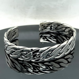 May include: A silver braided cuff bracelet with a smooth, polished finish. The bracelet is open and has a slightly curved shape.
