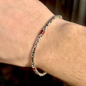 May include: A silver bracelet with a detailed chain design and small red accents. The bracelet is worn on a wrist, showcasing its intricate pattern and color contrast. The jewelry piece is a stylish accessory.