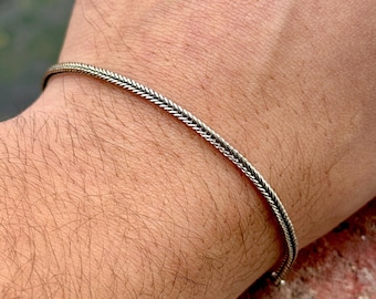 Könnte beinhalten: Ein silbernes Armband mit geflochtenem Design. Das Armband befindet sich an einem Handgelenk.