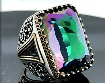 May include: A silver ring with a large, rectangular, iridescent gemstone. The gemstone displays a spectrum of colors, including green, purple, and blue. The ring has intricate filigree detailing on the sides and a gold-colored trim around the gemstone.