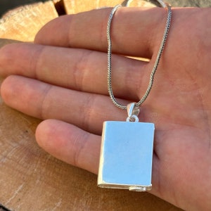 May include: A silver chain necklace with a silver locket pendant shaped like a book. The locket is closed and has a smooth, polished surface.