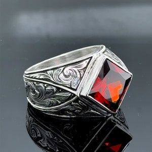 Può includere: Un anello d'argento con un intricato design floreale e una grande pietra rossa quadrata.