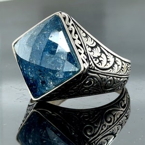 Op de afbeelding: Een zilveren ring met een grote, vierkante, blauwe edelsteen gezet in een gedetailleerd, versierd ontwerp.