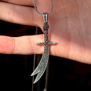 May include: A silver necklace featuring a detailed silver pendant shaped like a scimitar with red gemstones. The pendant has intricate designs and a pointed tip.
