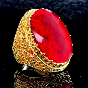 Op de afbeelding: Een gouden ring met een grote, rode, gefacetteerde edelsteen. De ring heeft een gedetailleerd, filigraan ontwerp.