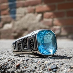 May include: A silver ring featuring a large, square, light blue gemstone. The ring's band has an ornate design. The ring is set on a textured concrete surface with a brick wall in the background.