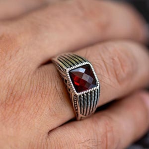 May include: A silver ring featuring a square, red gemstone. The ring's band has a textured design with vertical lines. The jewellery is worn on a finger, highlighting the ring's intricate details and the gemstone's colour.