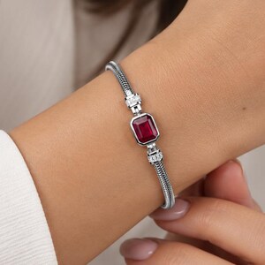 May include: A silver-toned bracelet featuring a central emerald-cut red gemstone. The bracelet has a flexible snake chain design with small, clear, round-cut stones set on either side of the central red stone, creating a refined and elegant look.