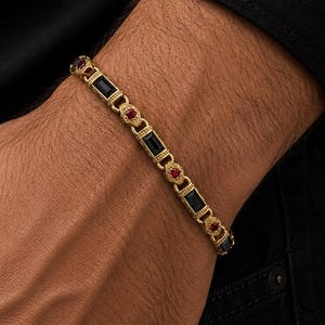 Op de afbeelding: Een goudkleurige schakelarmband met afwisselend rechthoekige zwarte stenen en kleine ronde rode stenen. De metalen schakels hebben een gestructureerd ontwerp met kralenranden, wat een herhalend patroon vormt over de lengte van het sieraad.