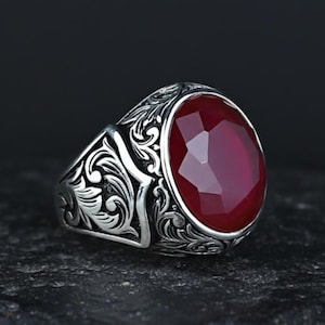 May include: A silver ring with an oval, faceted, deep red gemstone. The ring features intricate, swirling floral patterns on the band and around the gem. The gemstone is the focal point, catching the light and providing a striking contrast to the silver.