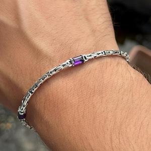 May include: A silver bracelet with a chain design, set with rectangular purple gemstones. The bracelet is worn on a wrist, highlighting the detailed craftsmanship. The purple gemstones provide a striking contrast to the silver.