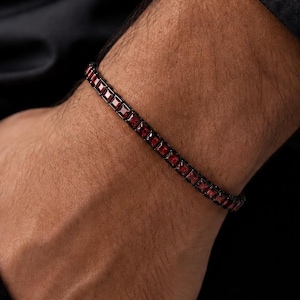 May include: A tennis bracelet featuring a series of square-cut, deep red gemstones set in a dark, black-toned metal frame. The bracelet consists of individual linked settings that hold each stone securely in a continuous, flexible line design.