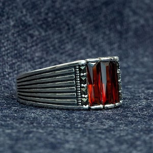 Op de afbeelding: Een zilveren ring met een rechthoekige rode edelsteen. De ring heeft een band met horizontale lijnen en kleine, donkere accentstenen. De ring wordt gepresenteerd op een donkerblauwe, getextureerde ondergrond.