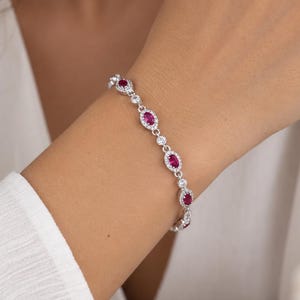 Può includere: Un bracciale color argento caratterizzato da una serie di pietre preziose ovali color rosa intenso. Ogni pietra è circondata da un alone di piccole pietre chiare e scintillanti. I collegamenti tra le montature ovali sono accentuati da singole pietre chiare.