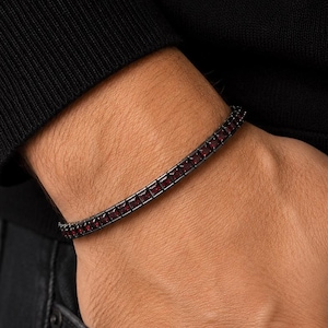 Könnte beinhalten: Ein Tennisarmband mit einer Reihe quadratisch geschliffener, tiefroter Edelsteine, die in einen dunklen, schwarzfarbenen Metallrahmen gefasst sind. Das Armband hat eine flexible Gliederstruktur und zeigt eine durchgehende Reihe von Steinen.