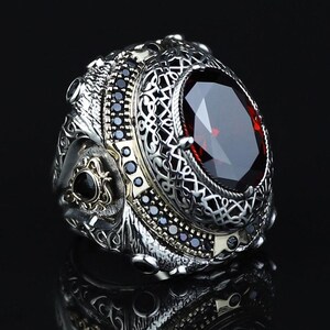 Puede incluir: Un anillo de plata con una piedra preciosa ovalada, facetada, de color rojo oscuro como pieza central. El anillo presenta intrincados filigranas y piedras negras alrededor de la piedra principal. El anillo tiene un diseño detallado y ornamentado, con un acabado pulido.