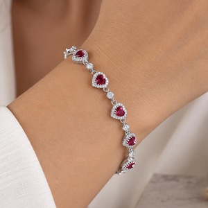 Op de afbeelding: Een zilverkleurige armband met een reeks hartvormige rode stenen, elk omringd door een halo van kleine heldere witte steentjes. De schakels tussen de hartvormige zettingen zijn geaccentueerd met enkele heldere witte steentjes.